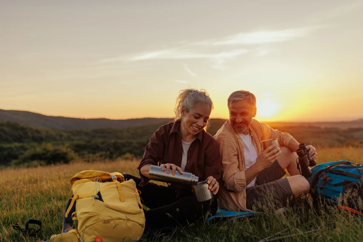 Middle age couple on outdoors hiking adventure. They are sitting at the foot of the mountain, woman pouring hot beverage into cups